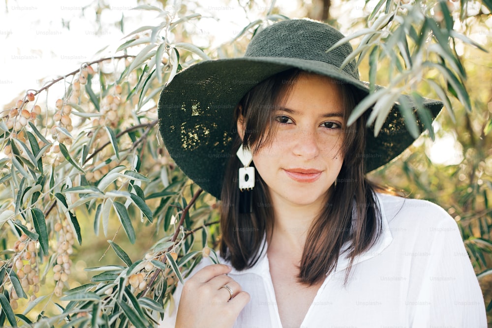 Retrato de una joven mujer de moda con pendientes modernos posando en ramas de olivo verde en suave luz de la tarde, elegante chica boho con sombrero relajándose en una isla tropical. Veraneo