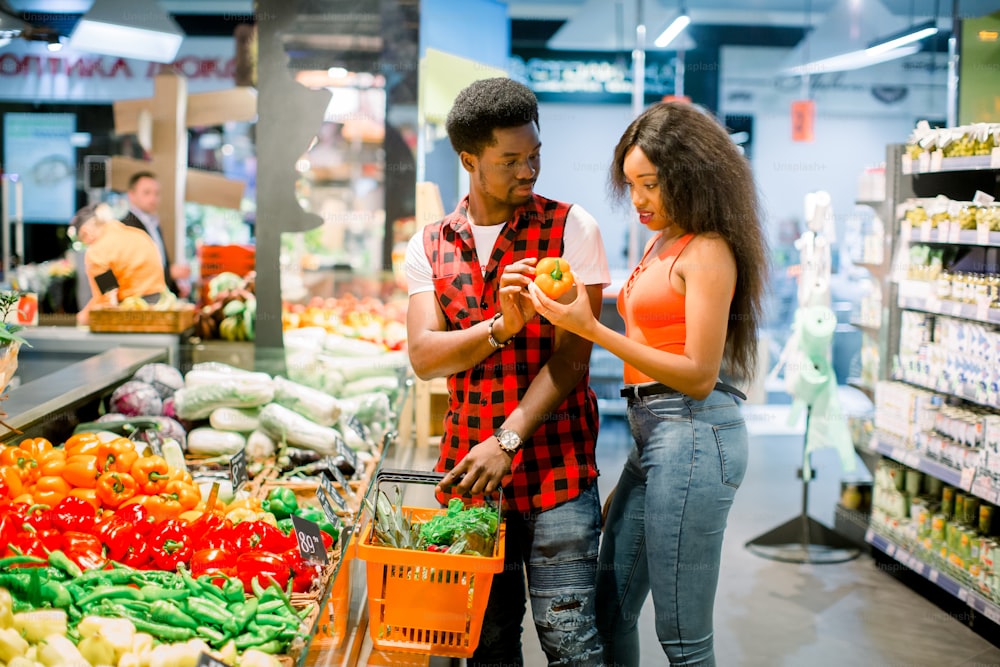 Joven pareja africana en ropa casual eligiendo verduras y frutas en el departamento de comestibles del supermercado, el hombre sostiene la cesta de la compra