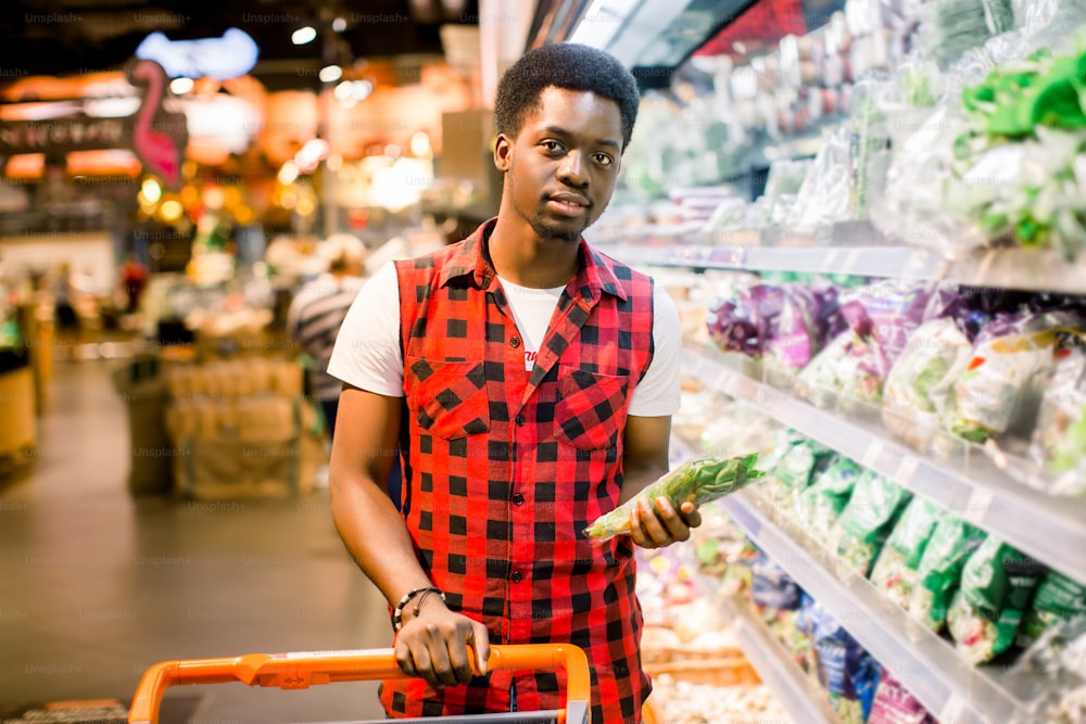 African man shopping in vegetable section at supermarket. Black man doing shopping at market while buying vegetables. Handsome guy holding shopping basket reading nutritional values of product.