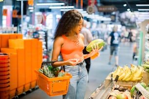 pretty african american woman in casual clothes with shopping basket in vegetable and fruit department in supermarket