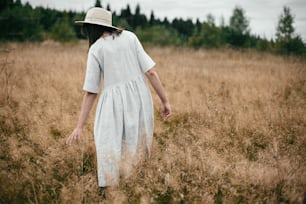 린넨 드레스와 모자를 쓴 세련된 소녀가 들판에서 허브와 야생화 사이를 걷고 있다. 시골에서 휴식을 취하는 보헤미안 여자, 단순한 느린 삶, 아미쉬 스타일. 텍스트를 위한 공간입니다. 대기 이미지