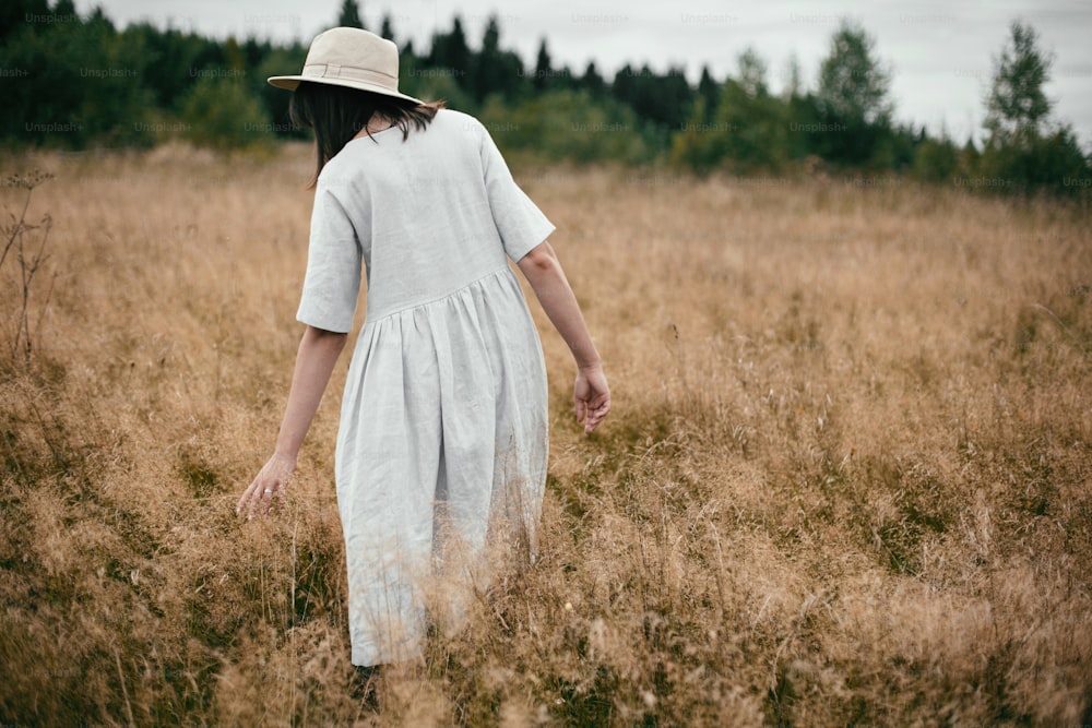 Stylish girl in linen dress and hat walking among herbs and wildflowers in field. Boho woman relaxing in countryside, simple slow life, amish style. Space for text. Atmospheric image