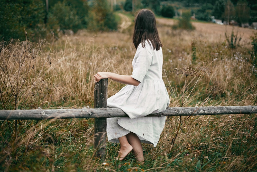 Stilvolles Mädchen im Leinenkleid, das auf einem alten Holzzaun zwischen Kräutern und Wildblumen sitzt und auf das Feld schaut. Boho-Frau, die sich auf dem Land entspannt, einfacher langsamer Lebensstil.  Atmosphärisches Bild