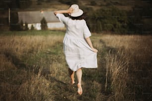 Stylish girl in linen dress running barefoot among herbs and wildflowers in sunny field in mountains. Boho woman relaxing in countryside, simple rustic life. Atmospheric image. Space text