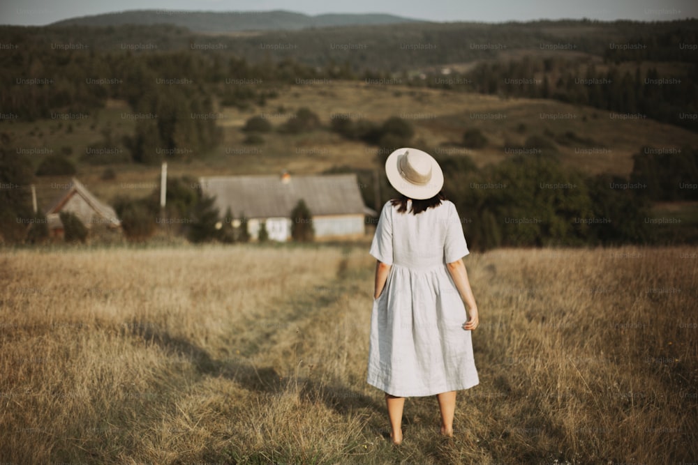 Stylish girl in linen dress and hat walking barefoot in grass in sunny field at village. Boho woman relaxing in countryside, simple rustic life. Atmospheric image. Space text