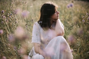 Stylish girl in rustic dress sitting among wildflowers and herbs in sunny meadow in mountains. Boho woman relaxing in countryside at sunset, simple life. Atmospheric image. Space text