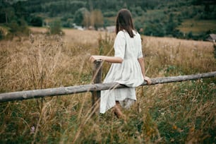 Fille élégante en robe de lin assise sur une clôture en bois vieillie parmi les herbes et les fleurs sauvages, regardant le champ. Femme bohème se relaxant à la campagne, style de vie simple et lent.  Image atmosphérique