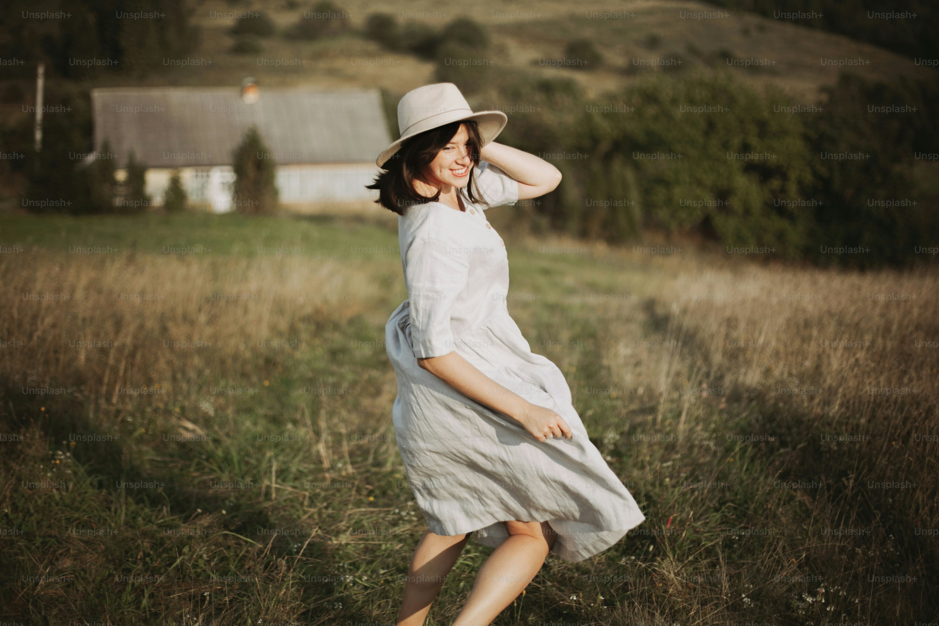 A woman in a light dress and hat walking through a grassy field