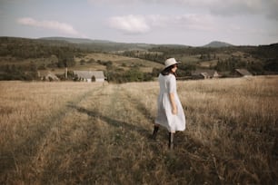 Muchacha elegante con vestido de lino y sombrero caminando entre hierbas y flores silvestres en un campo soleado en las montañas. Mujer boho relajándose en el campo, vida rústica sencilla. Imagen atmosférica. Texto espaciado