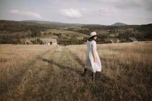 Stylish girl in linen dress and hat walking among herbs and wildflowers in sunny field in mountains. Boho woman relaxing in countryside, simple rustic life. Atmospheric image. Space text