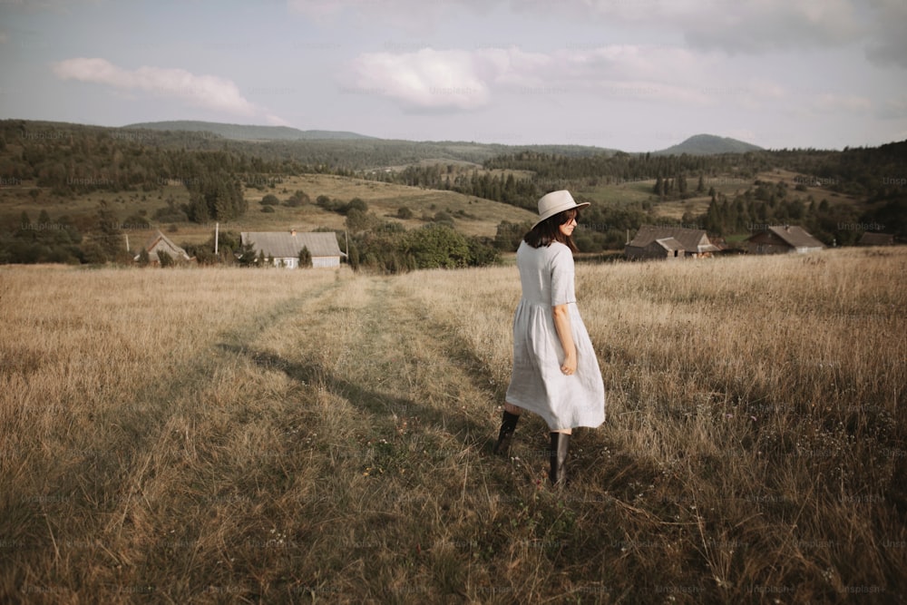 Stylish girl in linen dress and hat walking among herbs and wildflowers in sunny field in mountains. Boho woman relaxing in countryside, simple rustic life. Atmospheric image. Space text