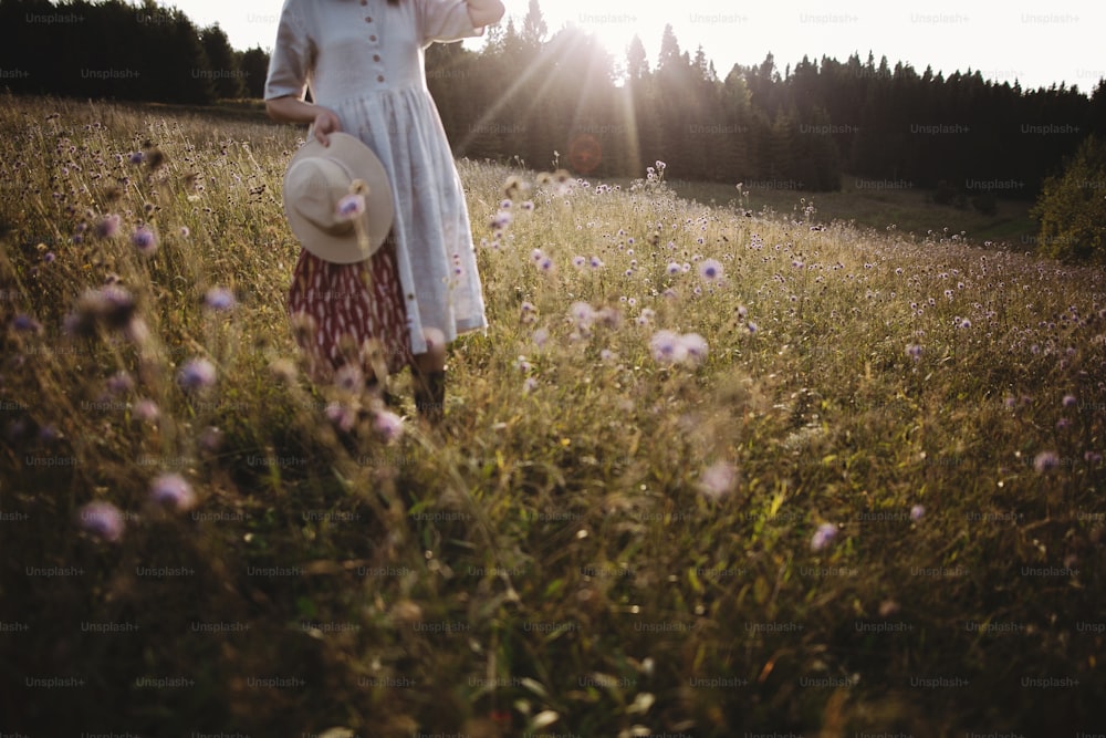 Wildblumen und Kräuter auf sonniger Wiese in Bergen und verschwommenes Bild eines stilvollen Mädchens in rustikalem Kleid und Hut. Boho Frau entspannt sich auf dem Land bei Sonnenuntergang, einfaches Leben. Atmosphärisches Bild