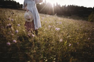 Wildflowers and herbs in sunny meadow in mountains and blurred image of stylish girl in rustic dress and hat. Boho woman relaxing in countryside at sunset, simple life. Atmospheric image