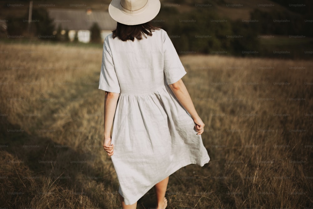 Stylish girl in linen dress and hat walking barefoot among herbs and wildflowers in sunny field in mountains. Boho woman relaxing in countryside, simple rustic life. Atmospheric image