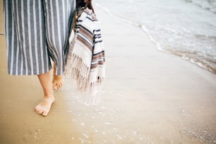 Stylish hipster girl walking barefoot on beach, holding bag  in hand, closeup. Summer vacation. Space for text. Calm moment. Boho woman relaxing at sea, enjoying walk on tropical island