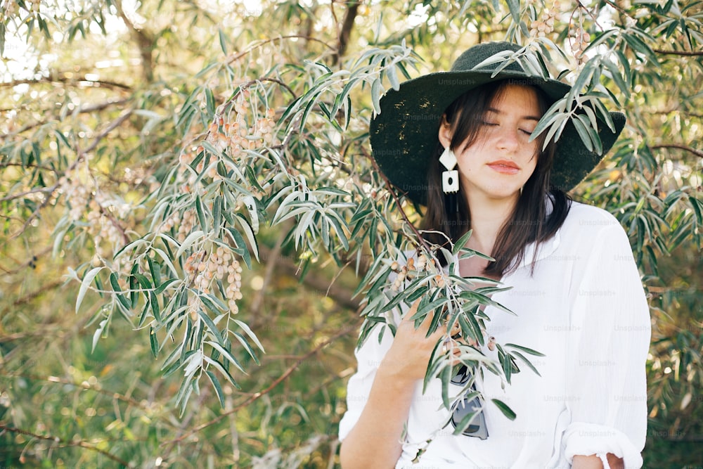 Retrato de una joven mujer de moda con pendientes modernos posando en ramas de olivo verde en la suave luz de la tarde, elegante chica boho relajándose en una isla tropical. Veraneo. Espacio para el texto