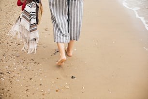 Chica hipster con estilo caminando descalza en la playa, sosteniendo bolso y zapatos en la mano, primer plano. Veraneo. Espacio para el texto. Momento de calma. Mujer boho relajándose en el mar, disfrutando de un paseo en una isla tropical