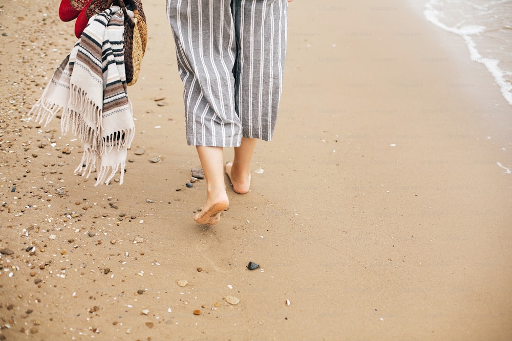 Stylish hipster girl walking barefoot on beach, holding bag and shoes in hand, closeup. Summer vacation. Space for text. Calm moment. Boho woman relaxing at sea, enjoying walk on tropical island