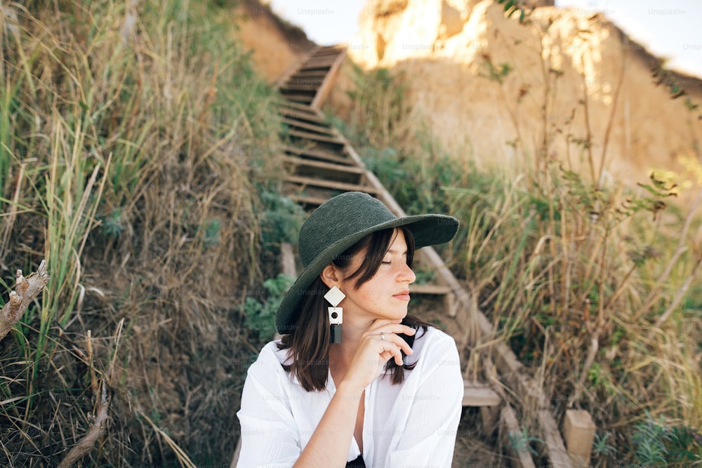 Elegante ragazza boho in cappello seduta su scale di legno in luce soleggiata sulla scogliera sabbiosa vicino al mare. Felice giovane donna alla moda che si rilassa sulla spiaggia dell'isola tropicale. Vacanze estive. Spazio per il testo