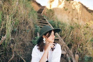 Stylish boho girl in hat sitting on wooden stairs in sunny light at sandy cliff near sea. Happy young fashionable woman relaxing on tropical island beach. Summer vacation. Space for text