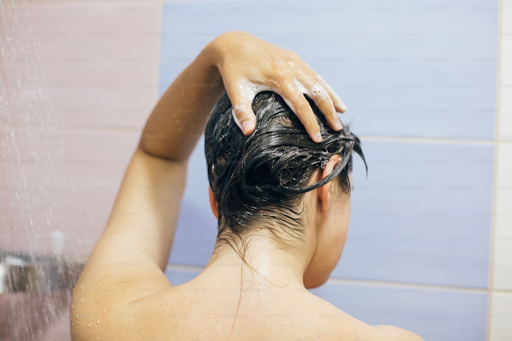 Junge glückliche Frau, die ihre Haare mit Shampoo wäscht, mit den Händen schäumt. Schönes brünettes Mädchen duschen und entspannende Zeit genießen. Körper-, Haar- und Hauthygiene, Lifestyle-Konzept.