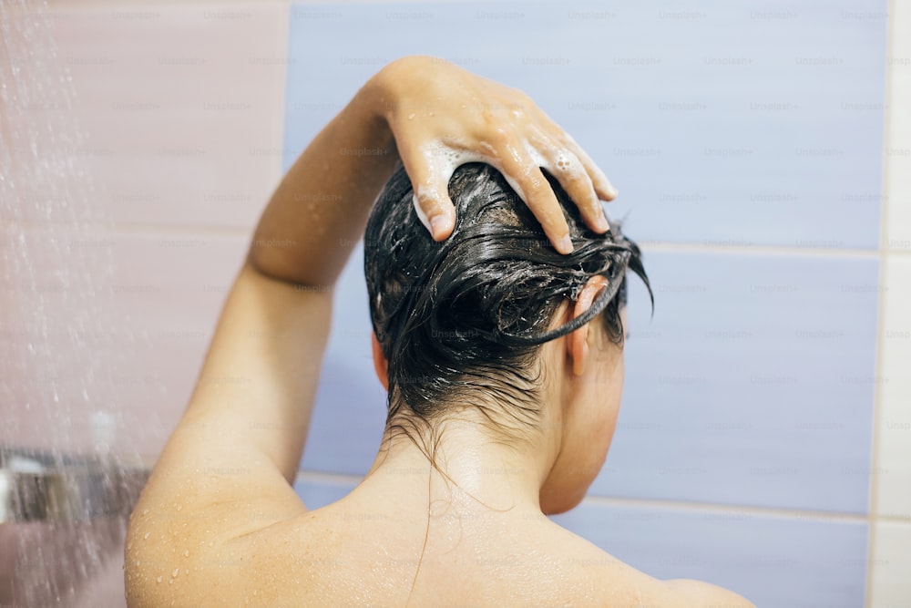 Young happy woman washing her hair with shampoo, foaming with hands. Beautiful  brunette girl taking shower and enjoying relax time. Body, hair and skin hygiene, lifestyle concept.