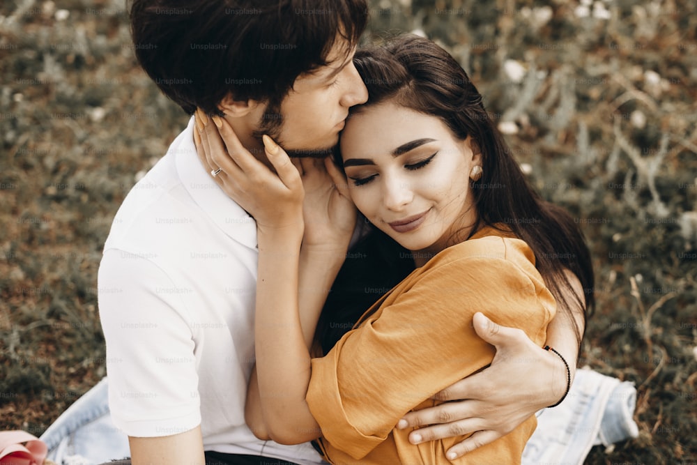 Close up portrait of a amazing caucasian woman with long dark hair leaning on her boyfriend with closed eyes smiling while sitting on field with flowers .