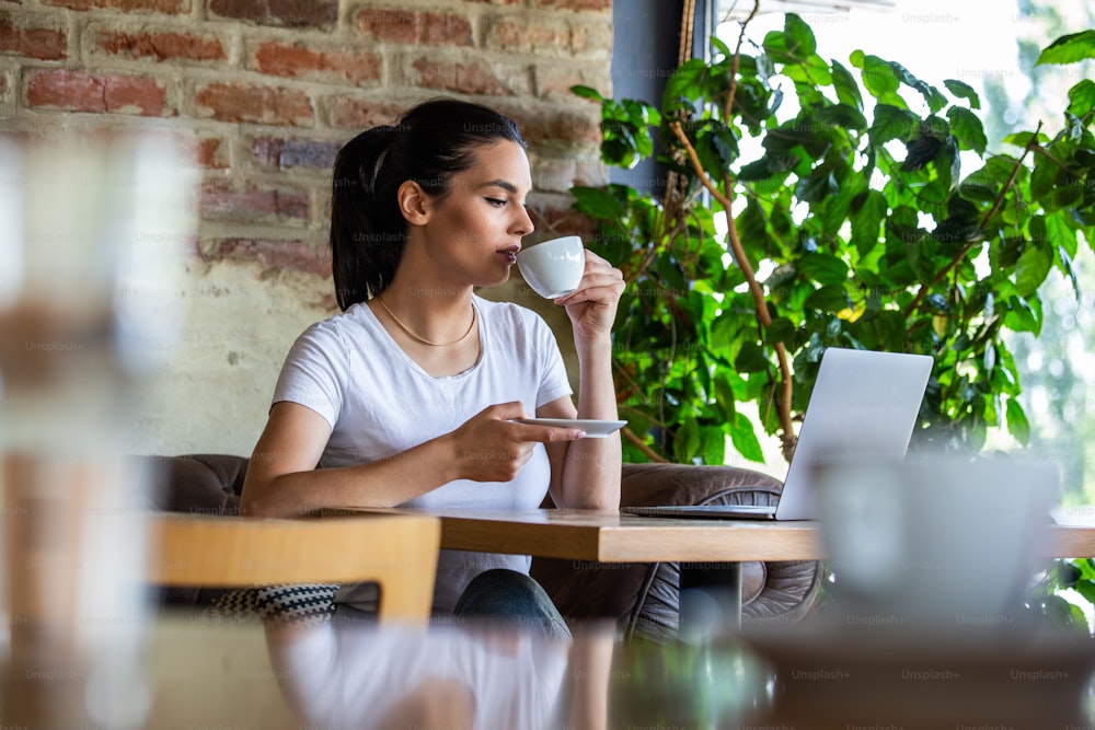 カフェに座ってコーヒーを飲みながら上機嫌の女性。明るく晴れた朝。コーヒーを飲みながら、コーヒーブレイクでネットサーフィンをするゴージャスな女性。朝のコーヒーは私の日課です
