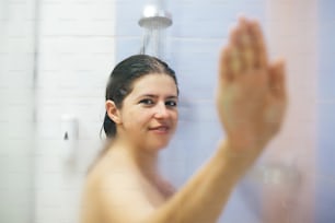 Young happy woman taking hot shower at home or hotel bathroom. Beautiful brunette girl holding hand on glass with steam, enjoying time in shower. Body and skin hygiene, lifestyle concept