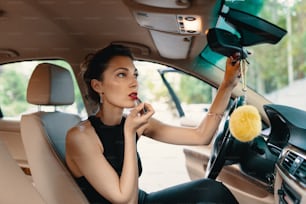 Giovane donna elegante che guarda nello specchietto retrovisore dipingendosi le labbra facendo applicare il trucco mentre guida l'auto.