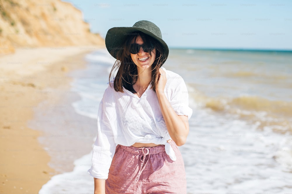 Happy young boho woman walking in sea waves in sunny warm day at tropical island and blue sky. Space for text. Stylish hipster girl in hat  relaxing on beach and smiling. Summer vacation