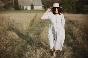 Stylish girl in linen dress and hat walking barefoot among herbs and wildflowers in sunny field in mountains. Boho woman smiling in countryside, simple rustic life. Atmospheric image