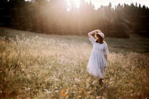 Stylish girl in rustic dress walking in wildflowers in sunny meadow in mountains. Boho woman relaxing in countryside flowers at sunset, rural simple life. Atmospheric image. Space text