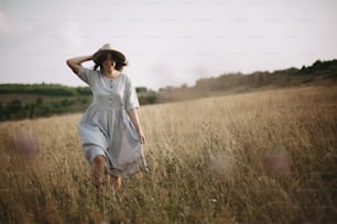 Stylish girl in linen dress running among herbs and wildflowers in sunny meadow in mountains. Boho woman relaxing in countryside, simple rustic life style. Atmospheric image. Space text