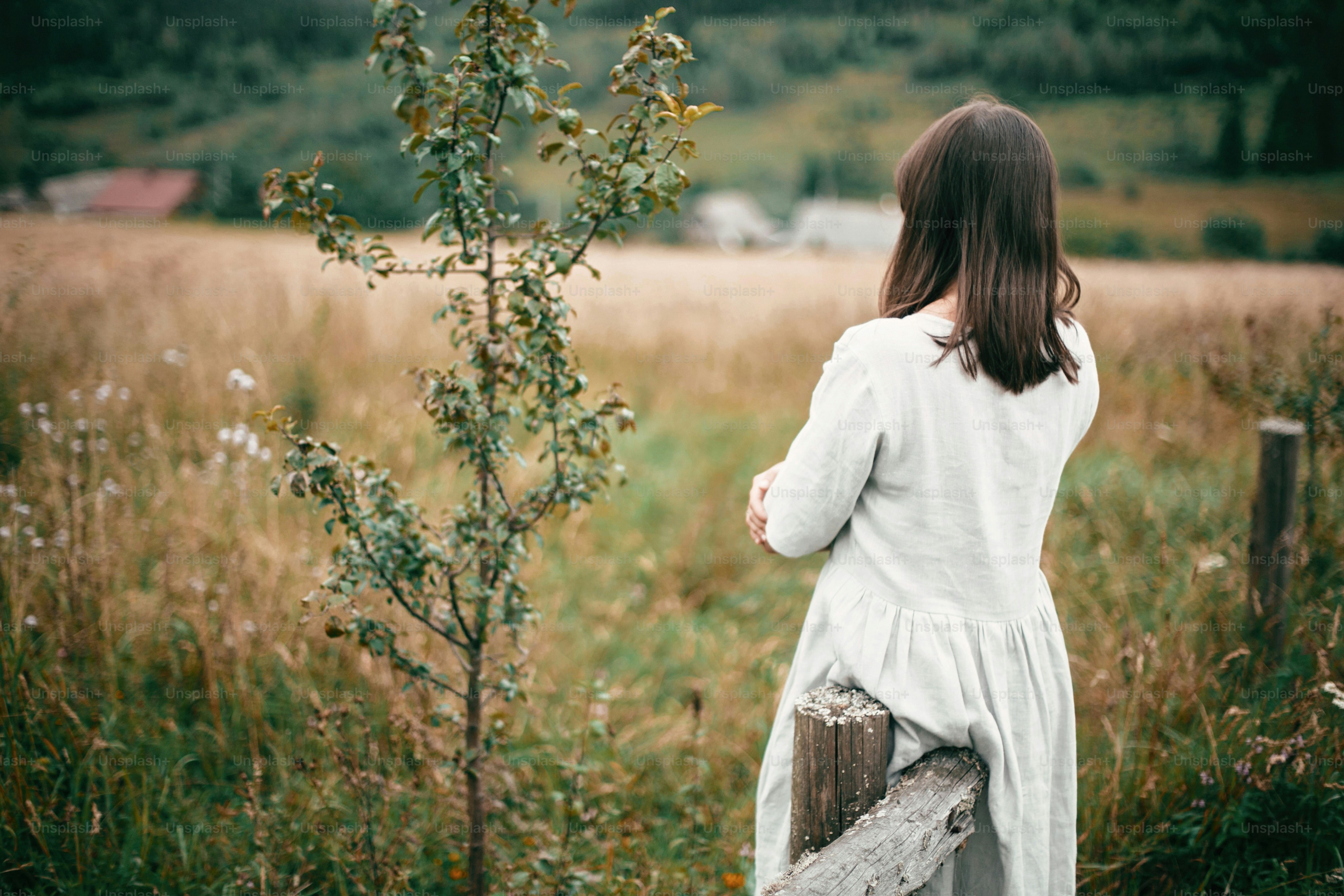 Stylish girl in linen dress standing at aged wooden fence among herbs and wildflowers, looking at field. Boho woman relaxing in countryside, simple slow life style.  Atmospheric image