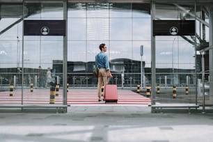 Ritratto a figura intera di ragazzo con borsa e in piedi vicino alle strisce pedonali vicino all'ingresso dell'aeroporto mentre distoglie lo sguardo. Copia spazio sul lato sinistro