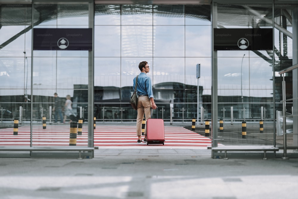 Retrato de corpo inteiro do cara com bolsa e de pé perto da faixa de pedestres perto da entrada do aeroporto enquanto desvia o olhar. Copiar espaço no lado esquerdo