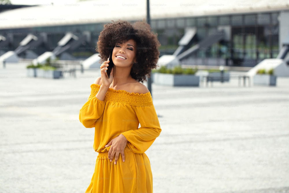 Portrait of young african american woman with curly hair posing in the city street, talking by mobile phone, smiling.