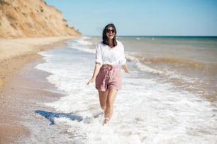 Summer vacation. Happy young boho woman walking and having fun in sea waves in sunny warm day on tropical island. Space for text. Stylish hipster girl relaxing on beach and smiling.
