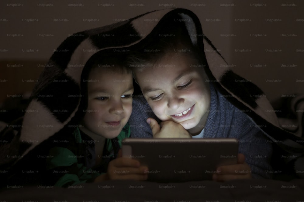 Two kids using tablet pc under blanket at night. Cute Brothers with tablet computer in a dark room smiling. Too entertained to sleep