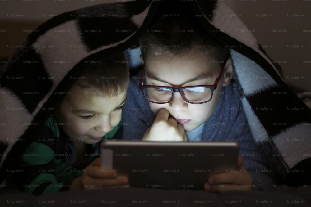 Due bambini che utilizzano tablet pc sotto coperta di notte. Fratelli carini con computer tablet in una stanza buia sorridente. Troppo divertito per dormire