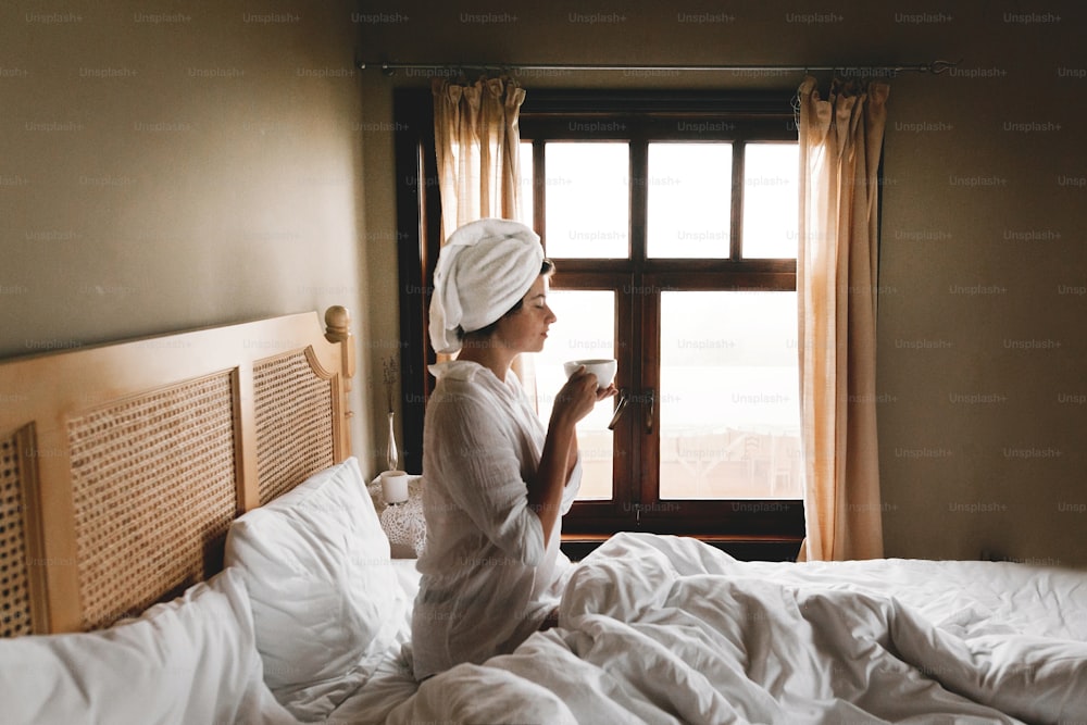 Beautiful happy young woman drinking coffee in bed in hotel room or home bedroom. Stylish brunette girl in white towel enjoying morning with hot drink. Space for text