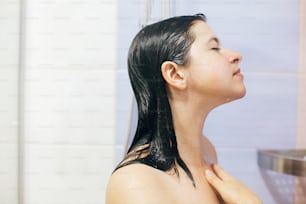 Young happy woman taking shower at home or hotel bathroom. Beautiful  brunette girl washing her hair and enjoying relax time. Body and skin hygiene, lifestyle concept