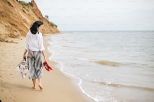 Stylish hipster girl walking barefoot on beach, holding bag and shoes in hand. Happy boho woman relaxing at sea, enjoying walk on tropical island. Summer vacation. Space for text.