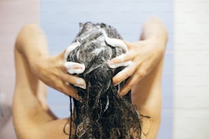 Young happy woman washing her hair with shampoo, hands with foam closeup. Back of beautiful brunette girl taking shower and enjoying relax time. Body and hair hygiene, lifestyle concept