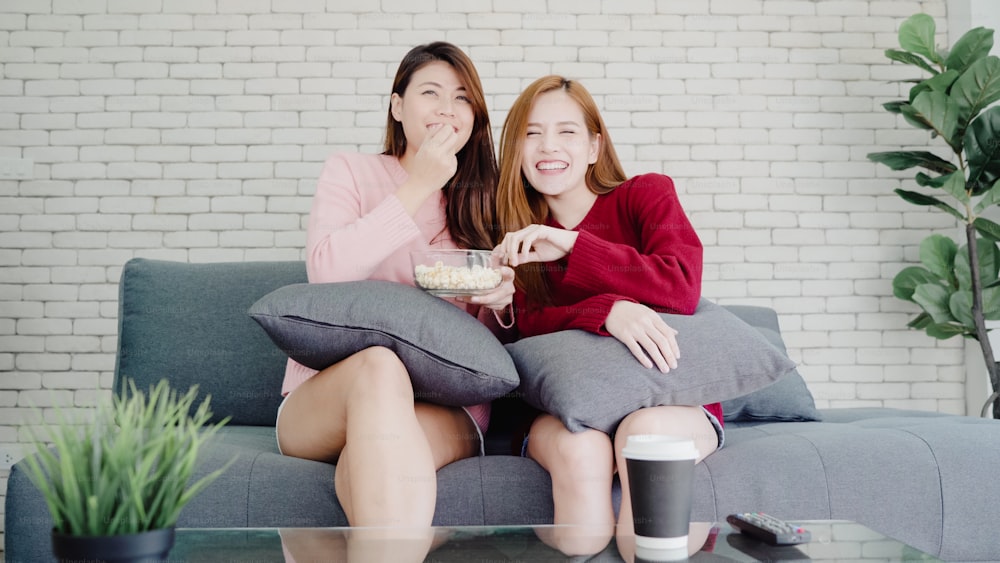 Lesbian Asian couple watching TV laugh and eating popcorn in living room at home, sweet couple enjoy funny moment while lying on the sofa when relaxed at home. Lifestyle couple relax at home concept.