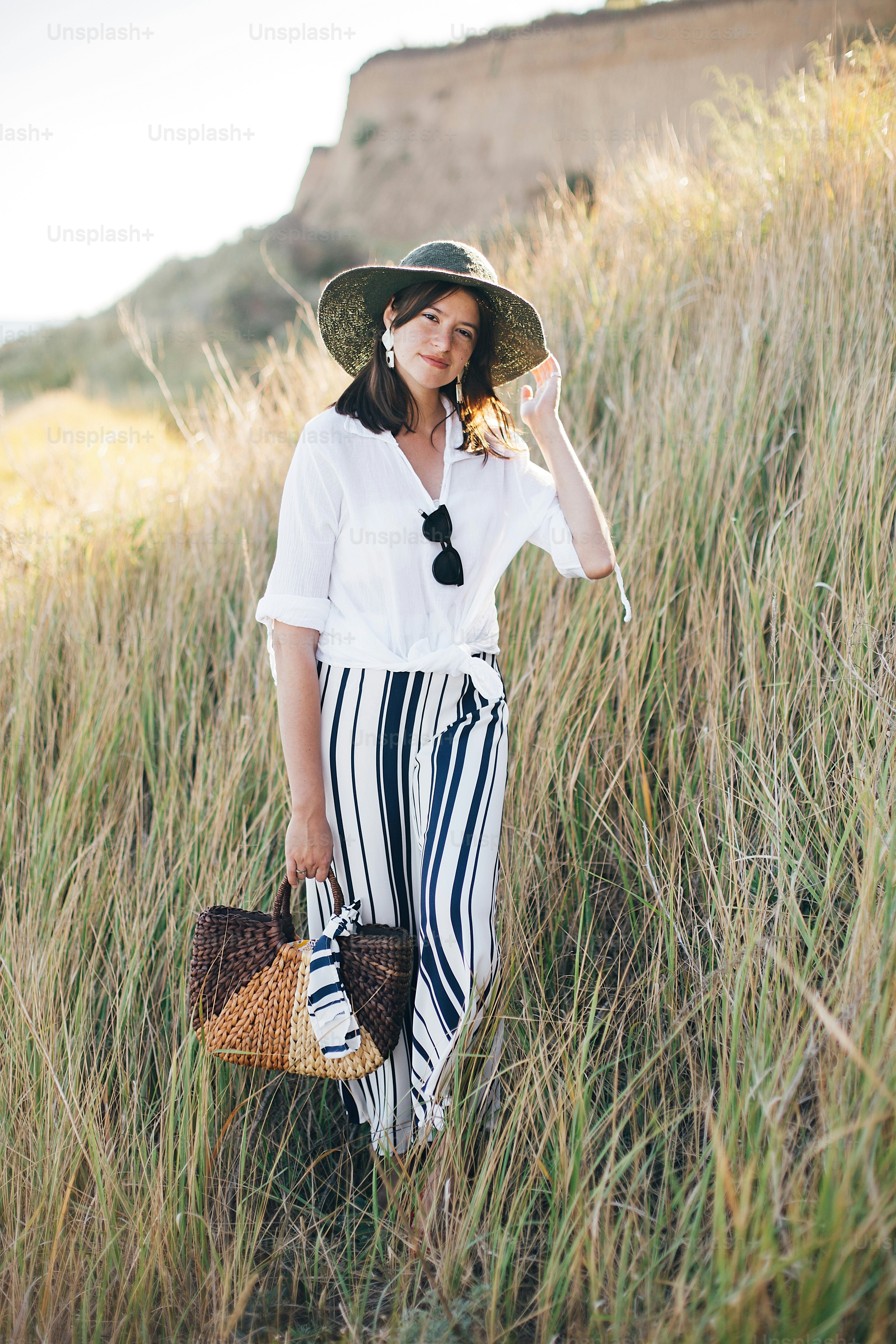 Stylish boho girl in hat posing among grass in sunny evening light at sandy cliff near sea. Happy young fashionable woman relaxing on tropical island beach. Summer vacation. Space for text