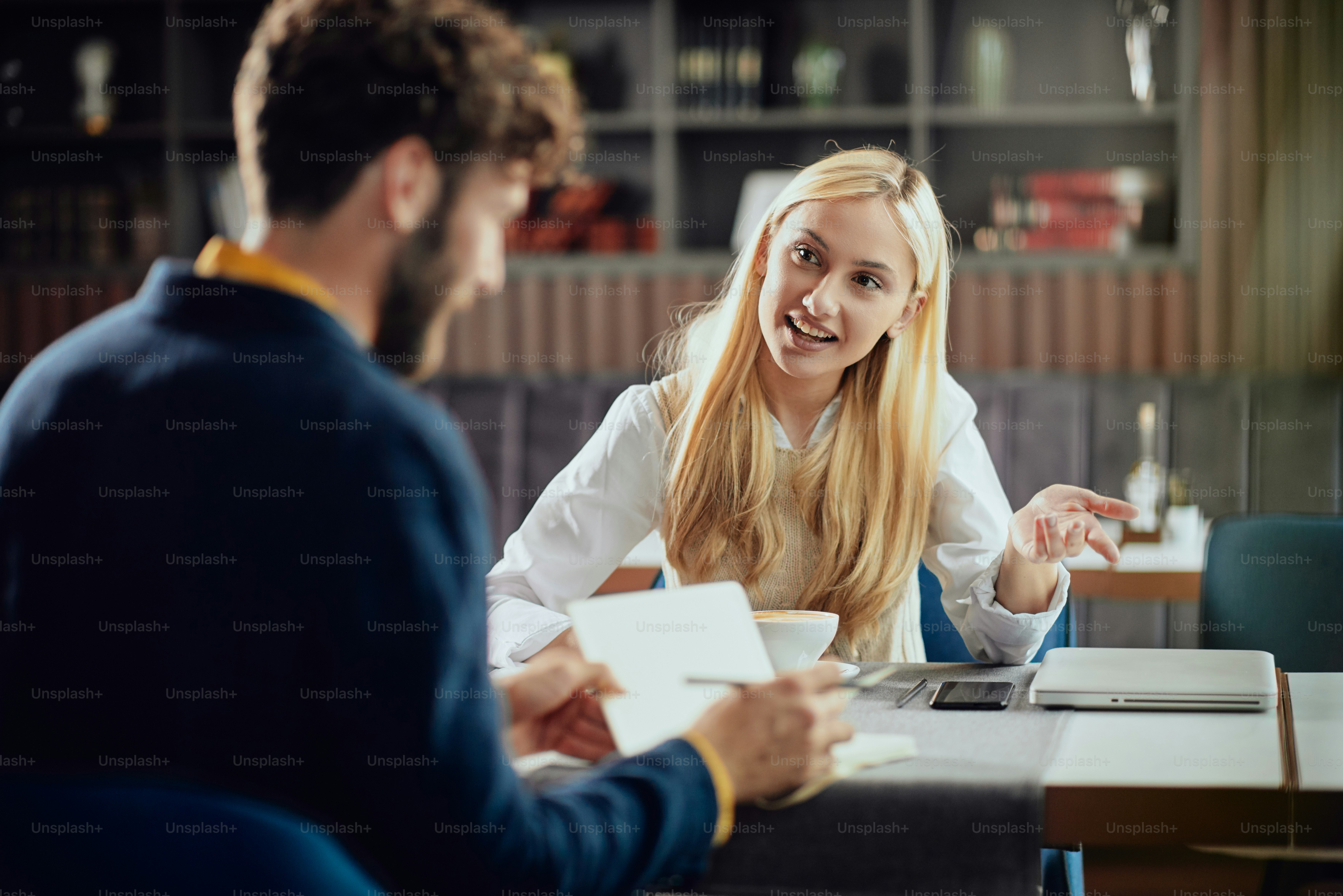 Smiling Caucasian blonde businesswoman dressed smart casual discussing with her male colleague about project while sitting in coffe shop.