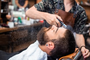 Homme barbu avec une longue barbe, brutal, hipster caucasien avec moustache, avec des cheveux élégants, coupe de cheveux, obtenir de la poudre sur la peau avec un pinceau de maquillage par le barbier ou les mains du coiffeur au salon de coiffure