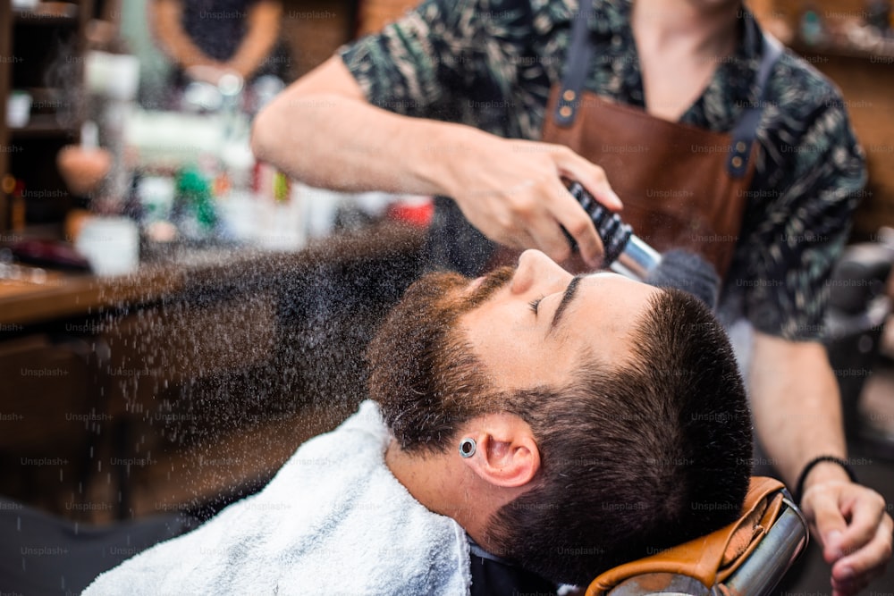 Bearded man with long beard, brutal, caucasian hipster with moustache, with stylish hair, haircut, getting powder on skin with makeup brush by barber or hairdresser hands at barbershop